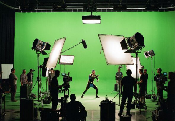 크고 밝은 녹색 배경 앞에 배우가 서 있고, 이를 비추는 다양한 영화 조명 장비가 배치된 전문적인 촬영